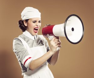 Portrait of angry young adult retro nurse screaming into megaphone.